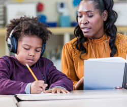 Student wears headphones and writes with pencil while teacher holds book and looks at student