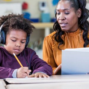 Student wears headphones and writes with pencil while teacher holds book and looks at student